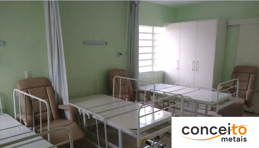 Enfermaria coletiva com múltiplas camas hospitalares articuladas, cada uma com grades e poltrona de acompanhante ao l...