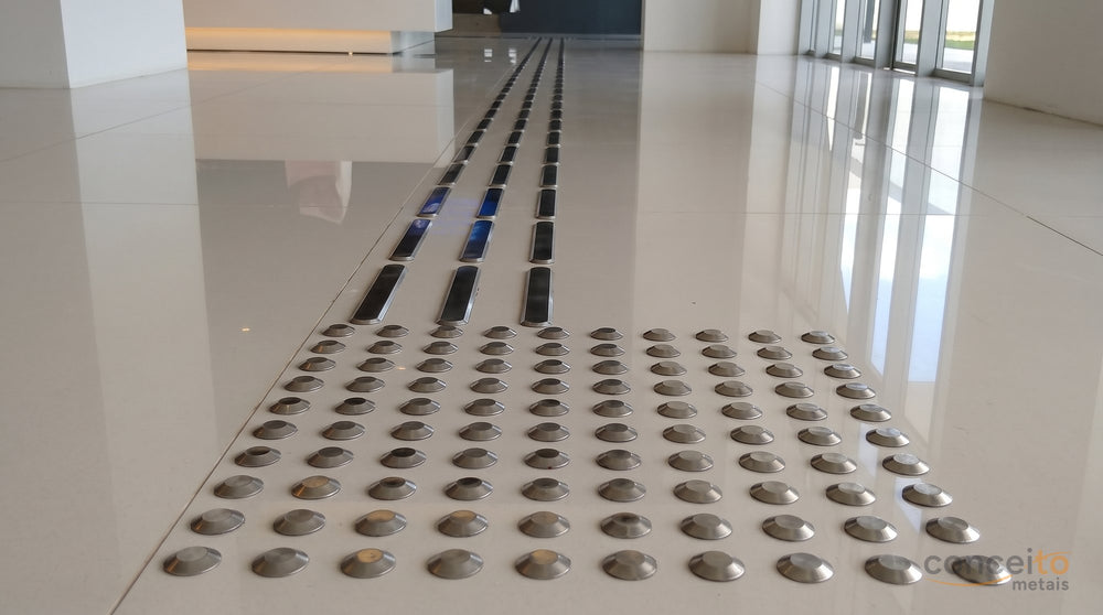 Piso tátil de alerta, com peças em relevo de botões, sinalizando uma mudança de piso ou obstáculo sobre o piso branco...