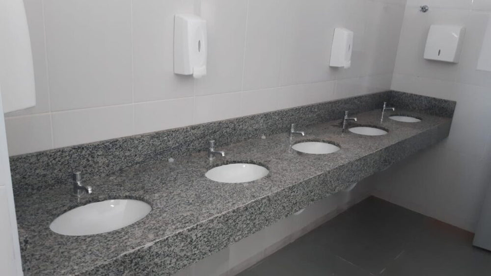 Bancada coletiva em granito com cinco cubas de lavatório, torneiras cromadas de acionamento individual e dispensadore...