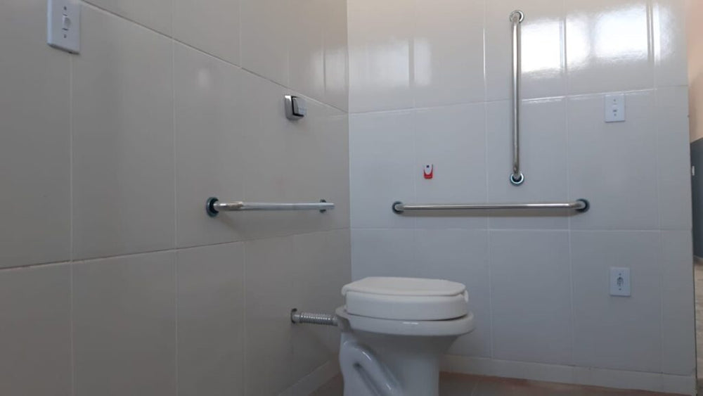 Banheiro PNE com vaso sanitário de assento elevado, barra de apoio reta e barra em L instaladas em inox conforme a NB...