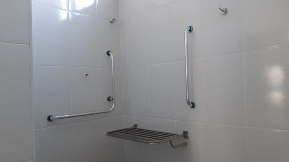 Box adaptado com barra de apoio reta vertical e barra em L em inox, além de banco articulado retrátil na posição aber...